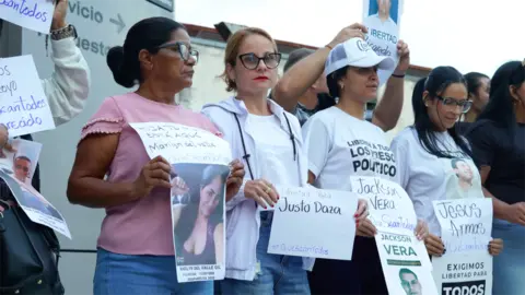 A group of women, most wearing T-shirts and jeans, stand outside El Helicoide prison holding signs 