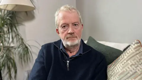 A man with short grey hair looks at the camera. He is seated, with cushions behind him and to his side. He is wearing a dark blue fleece with a zip, which is open at the neck