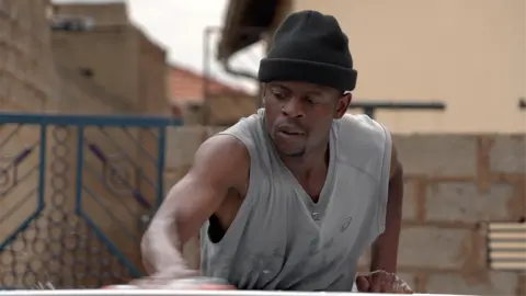 Christian Parkinson / BBC A shot of Arnold Ncube from the waist up. He is wearing a grey V-neck vest and black beanie hat. His right arm is slightly blurred as he can be seen wiping a car.