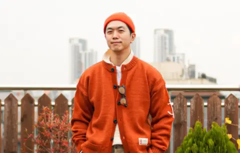 Instagram/@detailance Ji smiles at the camera with his hands in his pockets. He is wearing an orange sweater and orange beanie.