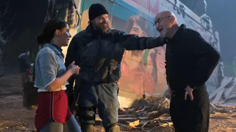 Netflix Millie Bobby Brown and David Harbour in character as Eleven and Jim Hopper. Eleven wears red shorts over grey leggings and a grey jumper. Jim Hopper wears a black leather jacket, a black beanie hat and dark trousers.They are speaking to director Frank Darabont on the set of Stranger Things. There is a vintage advertisement posted in the background with debrief on the ground.  