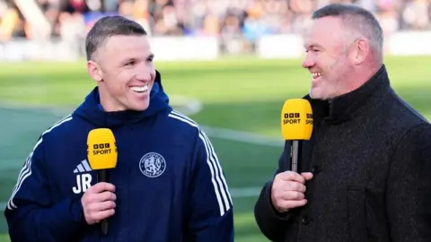 PA Media Two men are smiling while standing next to each other by the football pitch. They are both holding BBC Sport microphones.