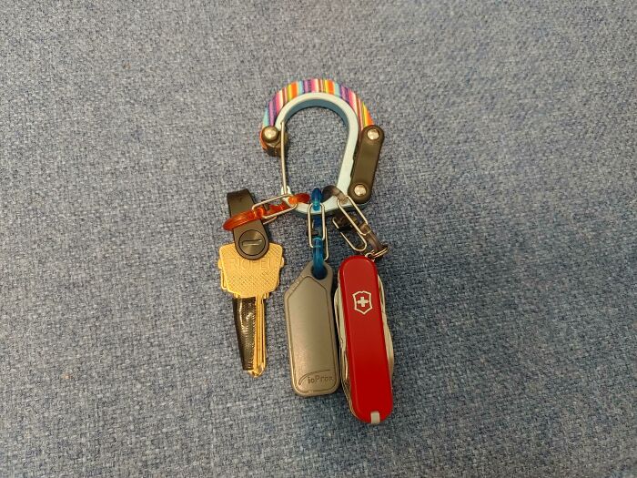 Colorful carabiner holding keys, a Swiss Army knife, and a Tile tracker on a blue fabric surface, symbolizing mysteries to solve.