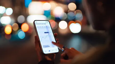 Getty Images Over the shoulder shot of somebody looking at financial performance on a smartphone