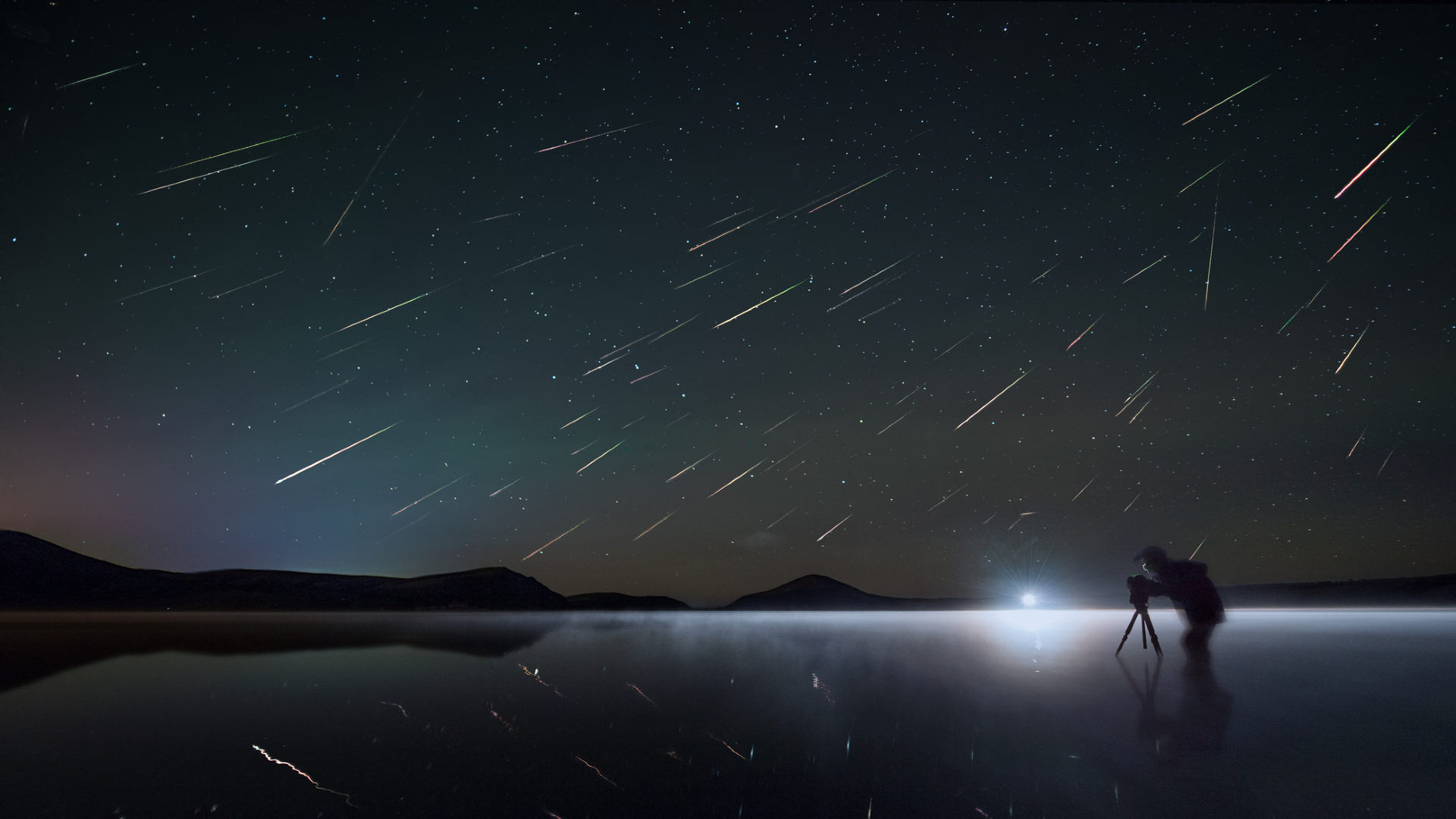 A figure with a tripod stands near a light illuminating a frozen lake with silhouettes of the mountains in the background with streaks of light filling the night sky above as the Perseid meteor shower gets underway