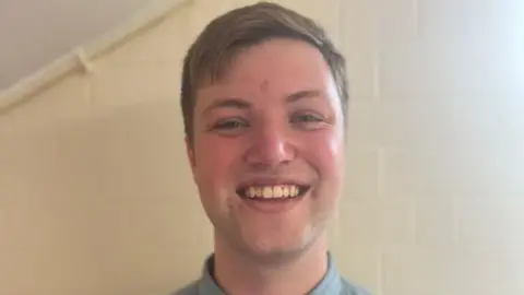 Conor Priestley Smiley young man, light brown hair, stands against a white wall. Conor.
