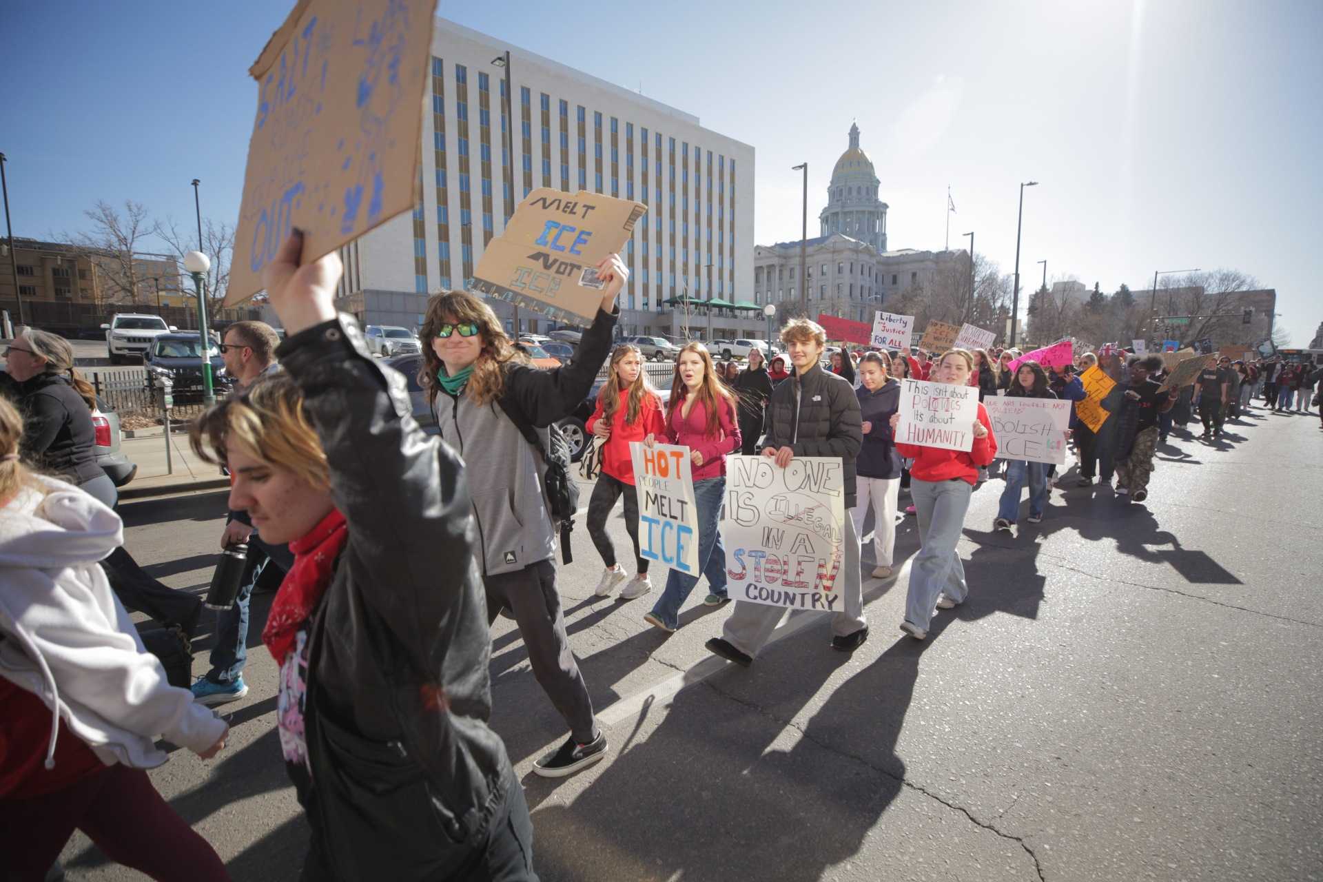 A large crowd of students walk in the street. Many wear red shirts or other articles of clothing. Many hold protest signs, but none are close enough to the camera to fully read in this photo.