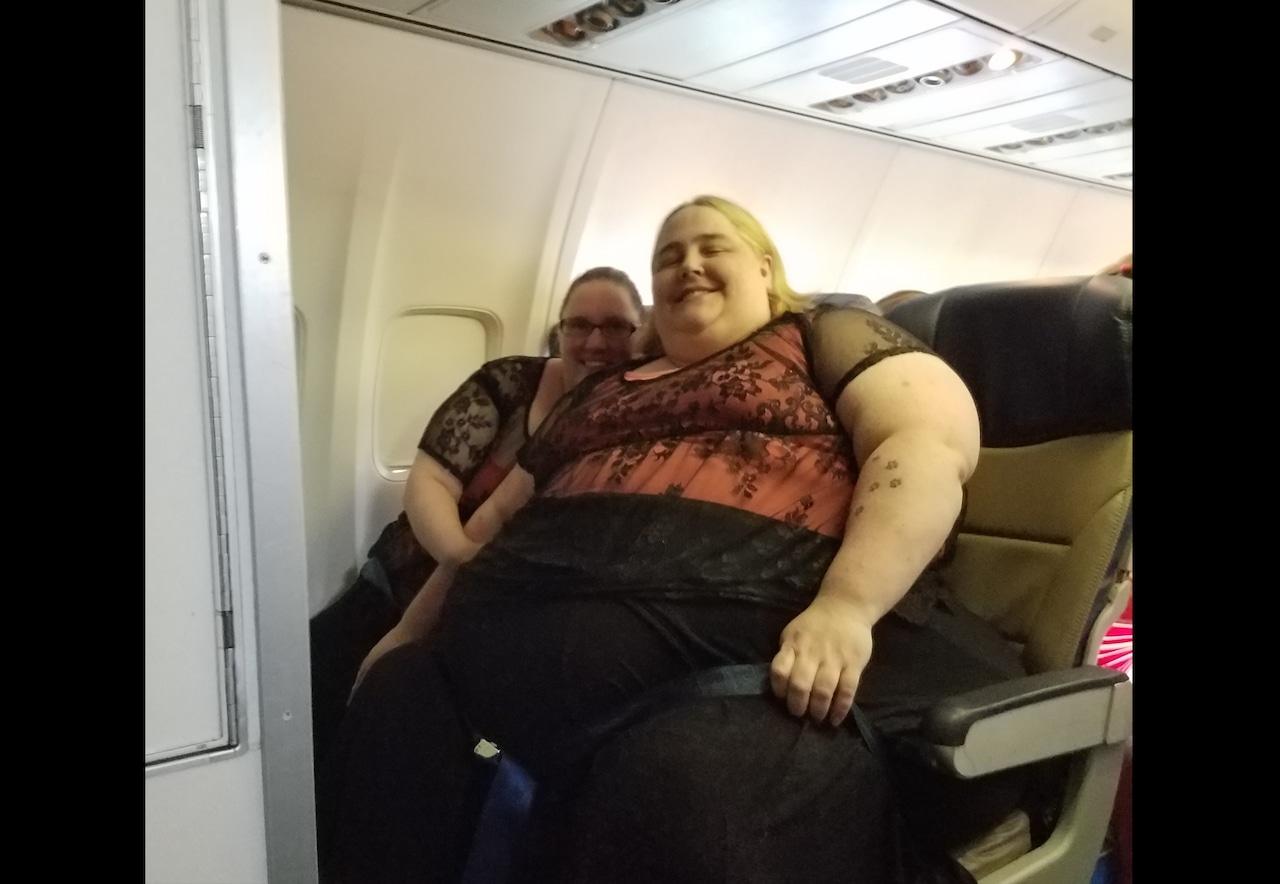 Two people sit in the bulkhead row of an airplane. One of them, a larger bodied person, takes up more than one of the seats.