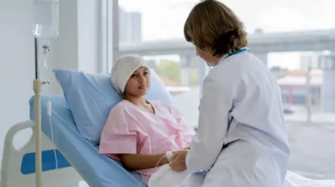 Getty Images A stock image of a patient wearing a pink hospital gown and head piece lies in a bed. A health professional is sitting on the bed holding her hand.