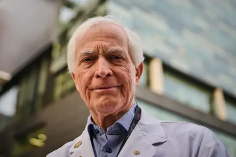 Dr Lang stands outside the neurology centre where he works, in Toronto, looking down at the camera in a white lab coat. His Order of Canada pin is on his lapel. 