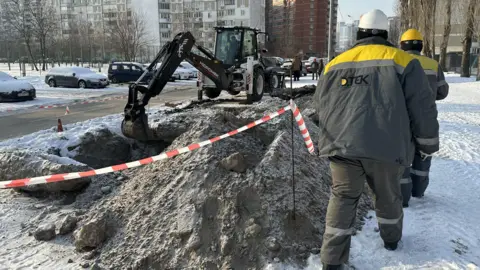 Two engineers in DTEK jackets walk away from the camera beside a pile of earth in the right of the picture as a digger excavates to their left beside a road