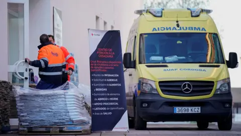 Reuters An ambulance and emergency workers outside the entrance to a civil building with a sign outside and what look like supplies. 