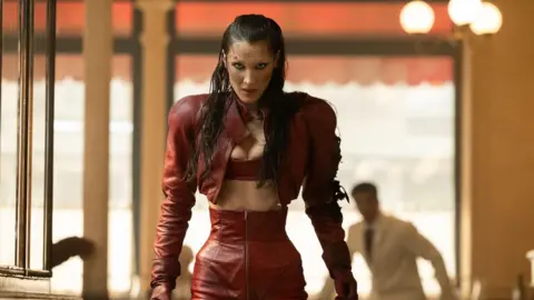 Disney via Getty Images Picture of Bella Hadid in a red outfit with a scary expression her face. The background is blurred but you can see a man in a white shirt.