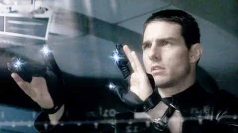 CBS Photo Archive Scene from the film Minority Report. A man (the actor Tom Cruise in the film Minority Report) stares at a transparent screen, wearing black gloves with bright lights on them. 