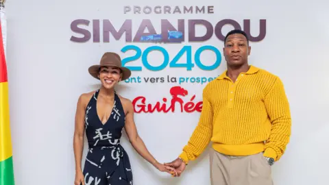 Mohamed Finando / Oxygen Africa Meagan Good (R) in a black-and-white jumpsuit and brown fedora, and Jonathan Majors (L) in a yellow jumper and beige slacks hold hands as they pose for the camera in front of a sign that reads: "Programme Simandou 2040." A Guinea flag hands furled on the far right of the frame
