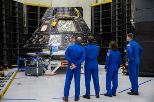 Four astronauts, wearing blue flight suits with their backs to the camera, look at the Orion spacecraft during the construction and testing process. 