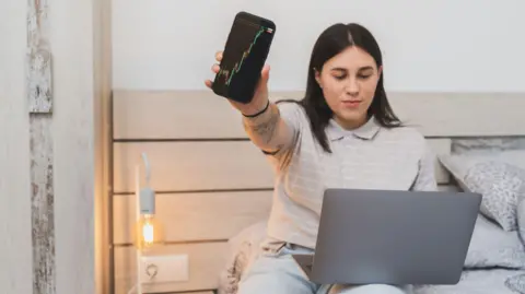 Getty Images Young woman sitting on a bed with a laptop on her legs and holding out a mobile phone with a graph on the screen.