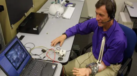 Kevin Warwick A man sitting down with a headset and wearing a purple shirt has his hand helf out in front of him, around his arm is a metal chip bracelet. He is looking at an open laptop screen.