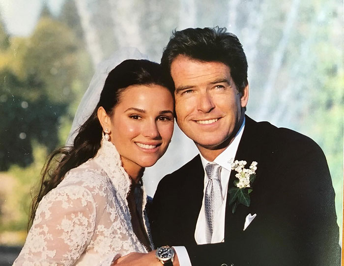 Pierce Brosnan with his wife on their wedding day, smiling and dressed in formal wedding attire outdoors.