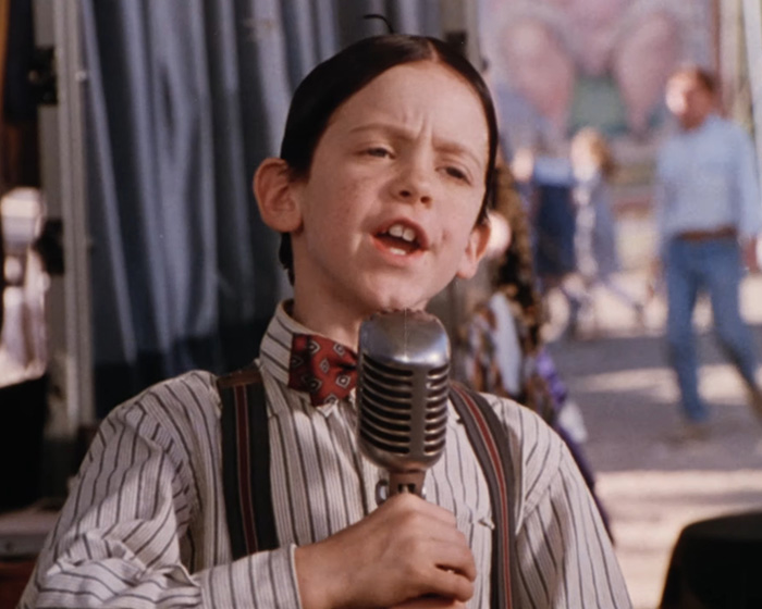 Child actor from Little Rascals speaking into a vintage microphone wearing a striped shirt and bow tie on set.