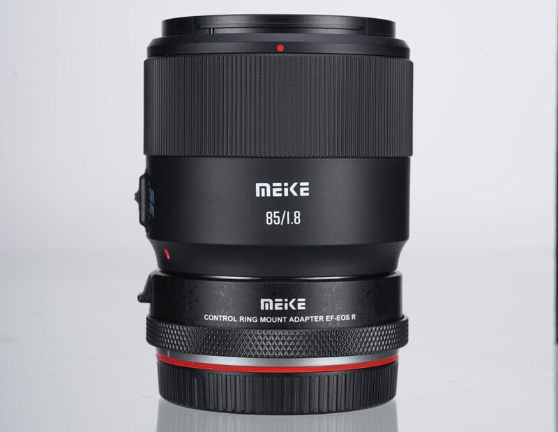 A Meike 85mm f/1.8 camera lens with a control ring mount adapter for EF-EOS R, shown standing upright on a white background. The lens and adapter have black bodies with white text and a red accent ring.