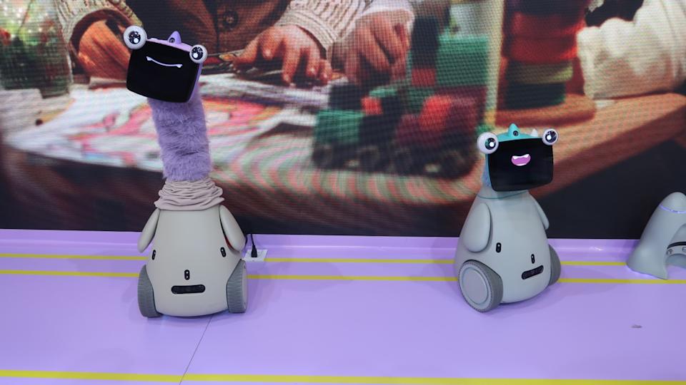 Two OlloBots — one with a long furry purple neck, making it about two feet taller than the other — are pictured on a light purple floor, in front of a screen displaying a closeup of a child playing with blocks.