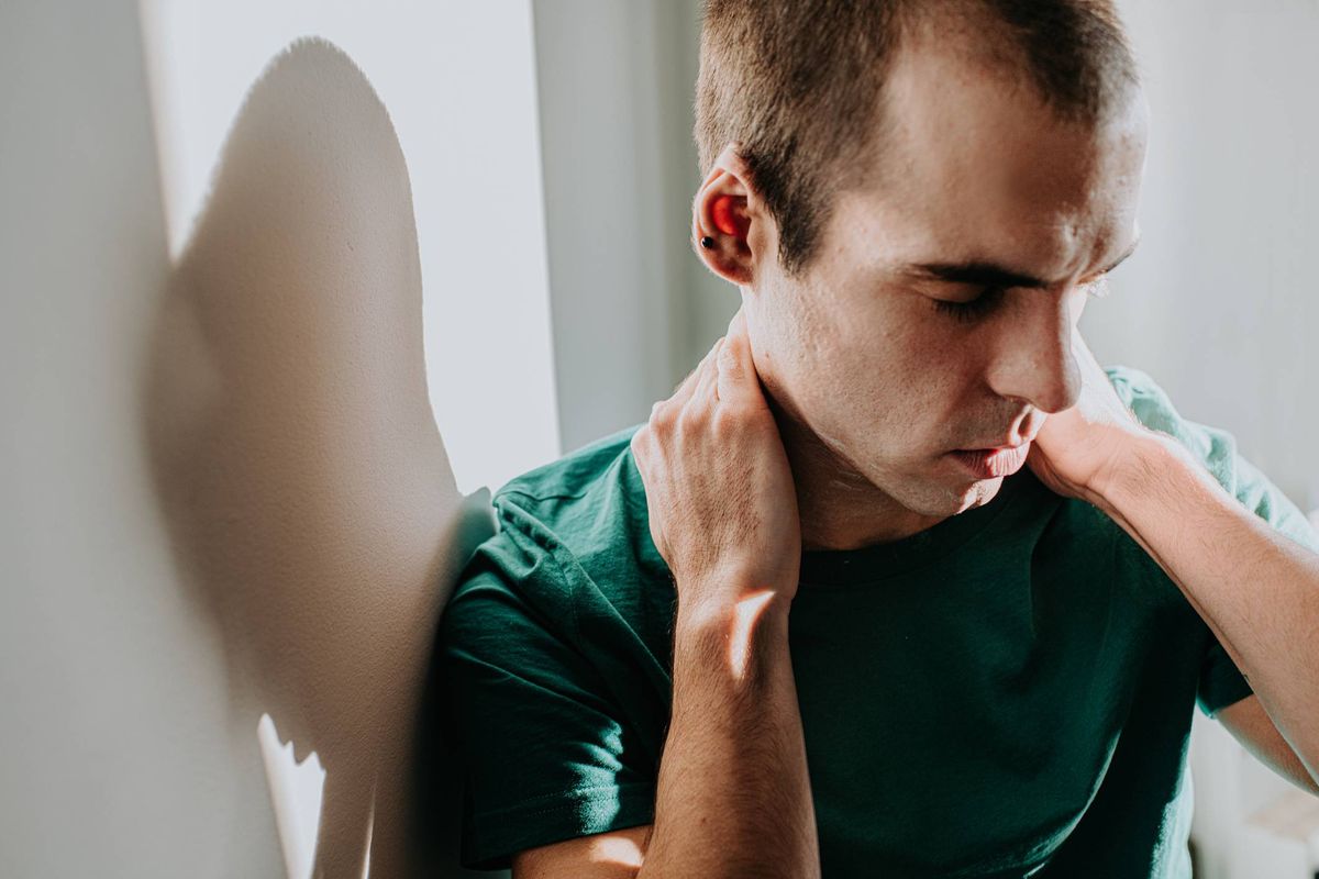Simple, conceptual image of a man rubbing his own neck with both hands. He looks serious, as if there is mounting emotional pressure, or a physical pain in his neck and shoulders. Wall provides space for copy.