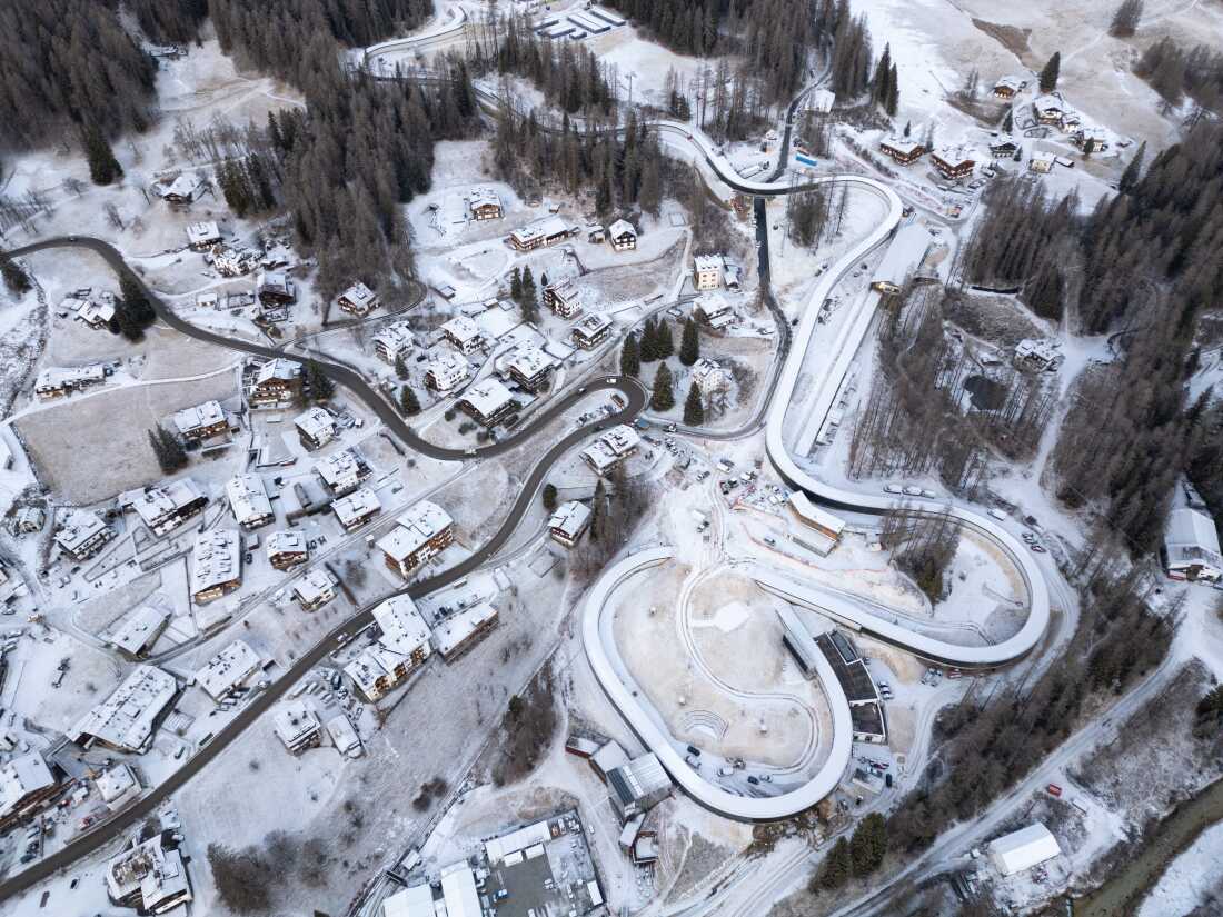 An overhead shot captures the construction of the new Cortina Sliding Centre, a track spanning approximately 1.4 kilometers with 16 curves, designed to host bobsleigh, skeleton, and luge events for the Milano Cortina 2026 Winter Olympics. The project has become a focal point for environmental protests after the felling of approximately 500 ancient larch trees in the Ronco forest to clear the path for the concrete structure