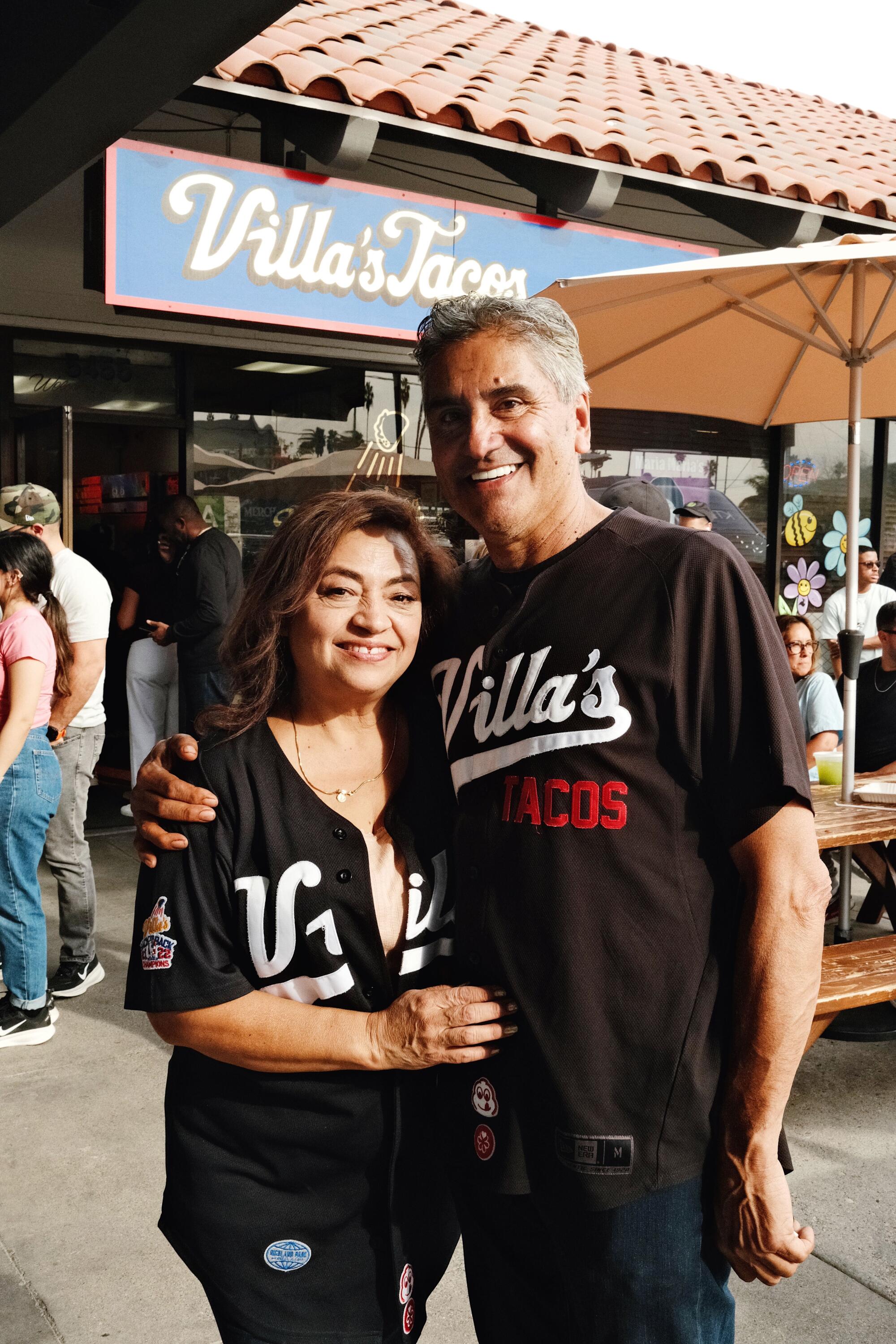 A man and woman wearing matching black baseball jerseys hug and smile at the camera