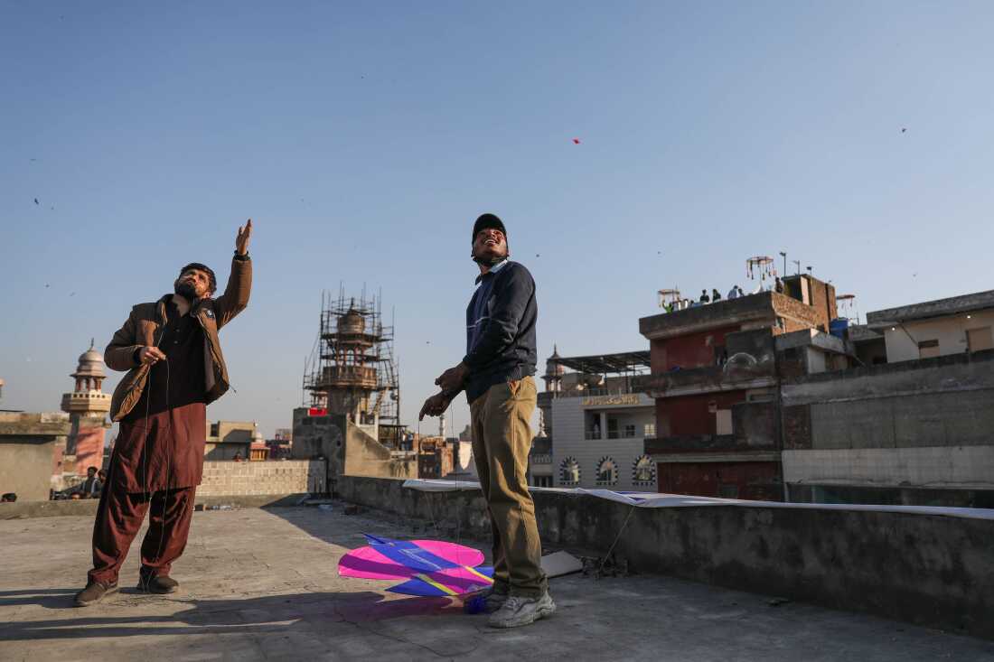 Two men stand on a rooftop and look at the sky as they fly kites. A building covered in scaffolding is scene behind them.