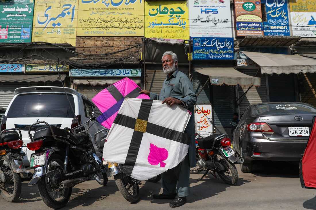 An older man with white hair and a white beard holds a pink kite in one hand and a white one in the other outside a market area near Mochi Gate during Basant. 