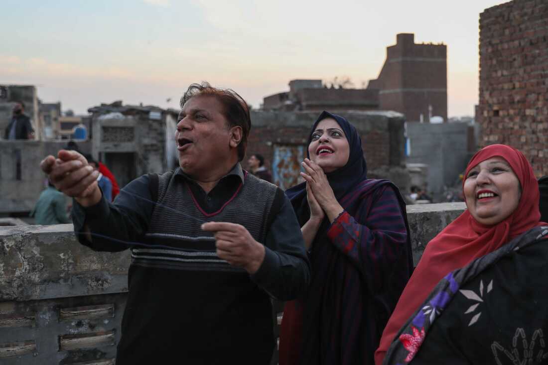 Zaib Un Nisa (center), who is wearing a black hijab and a patterned top, is smiling while watching her husband, Aqeel Ahmad, who is wearing a sweater vest over a dark shirt, fly a kite on a rooftop. He is also smiling and there is another woman wearing a red hijab standing nearby who is enjoying the kites.