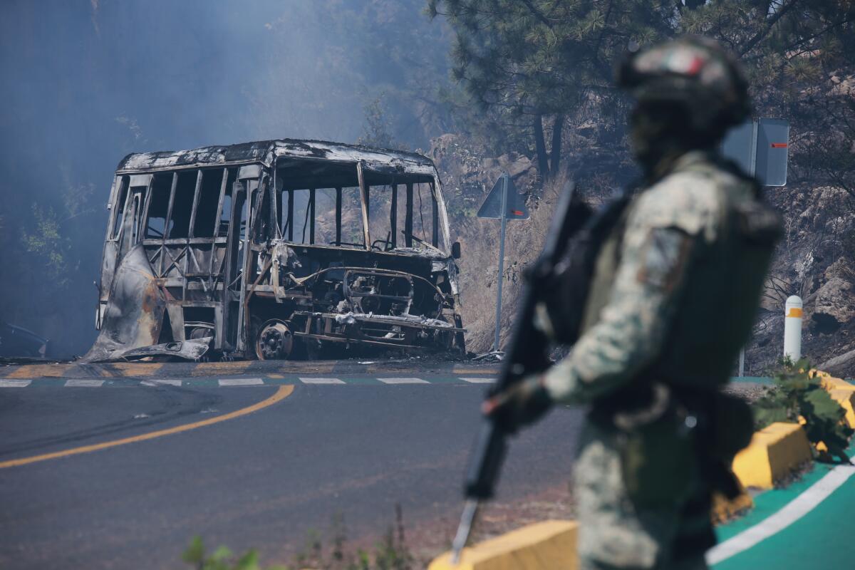 A soldier holding a weapon stands guard by a charred vehicle on a road