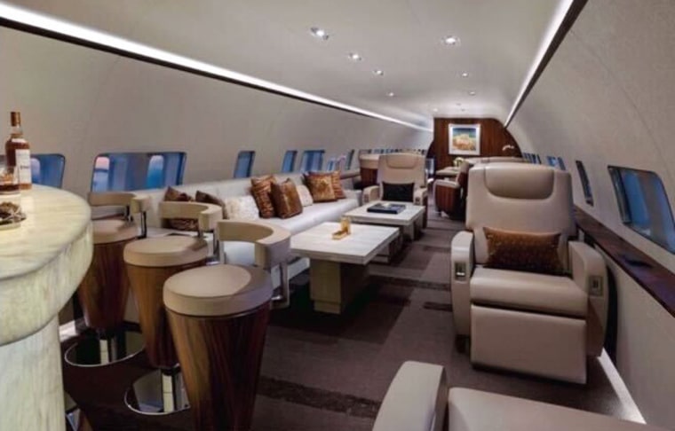The interior of a plane, beige reclining seats and bar stools can be seen