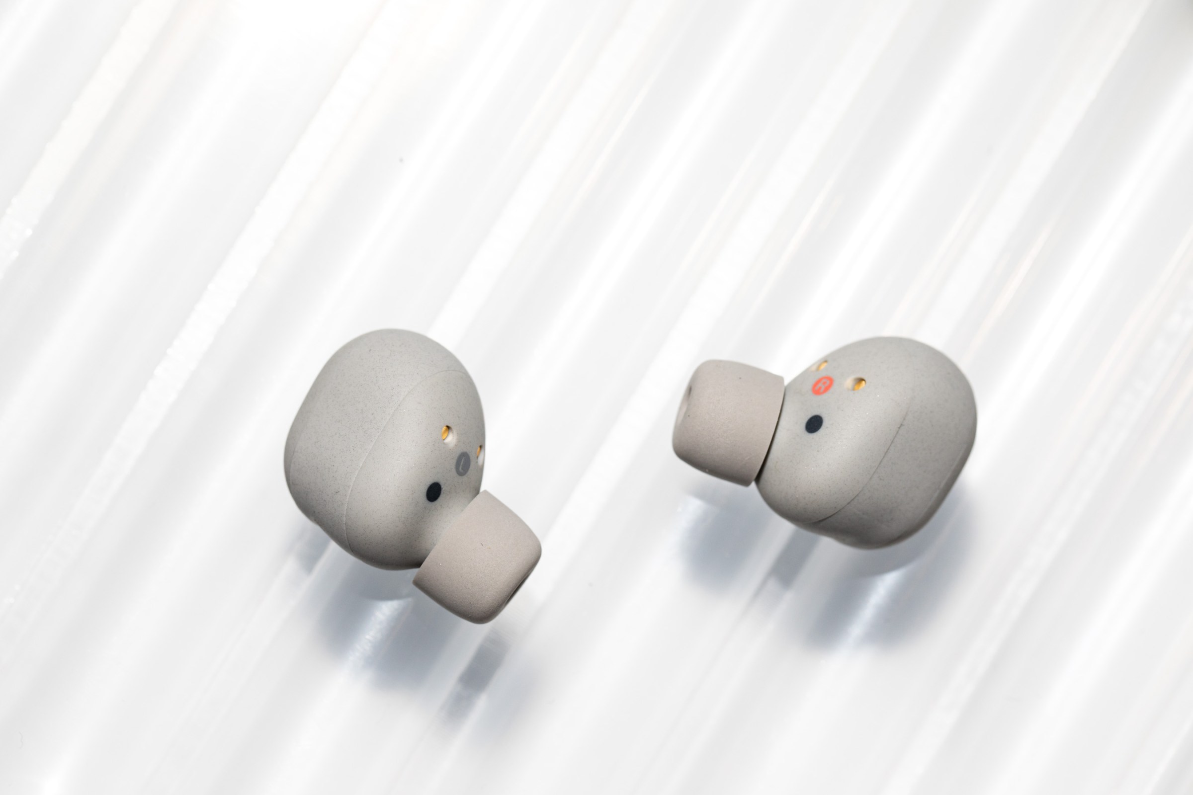 The Sony WF-1000XM6 earbuds in light gray on an off-white textured background.