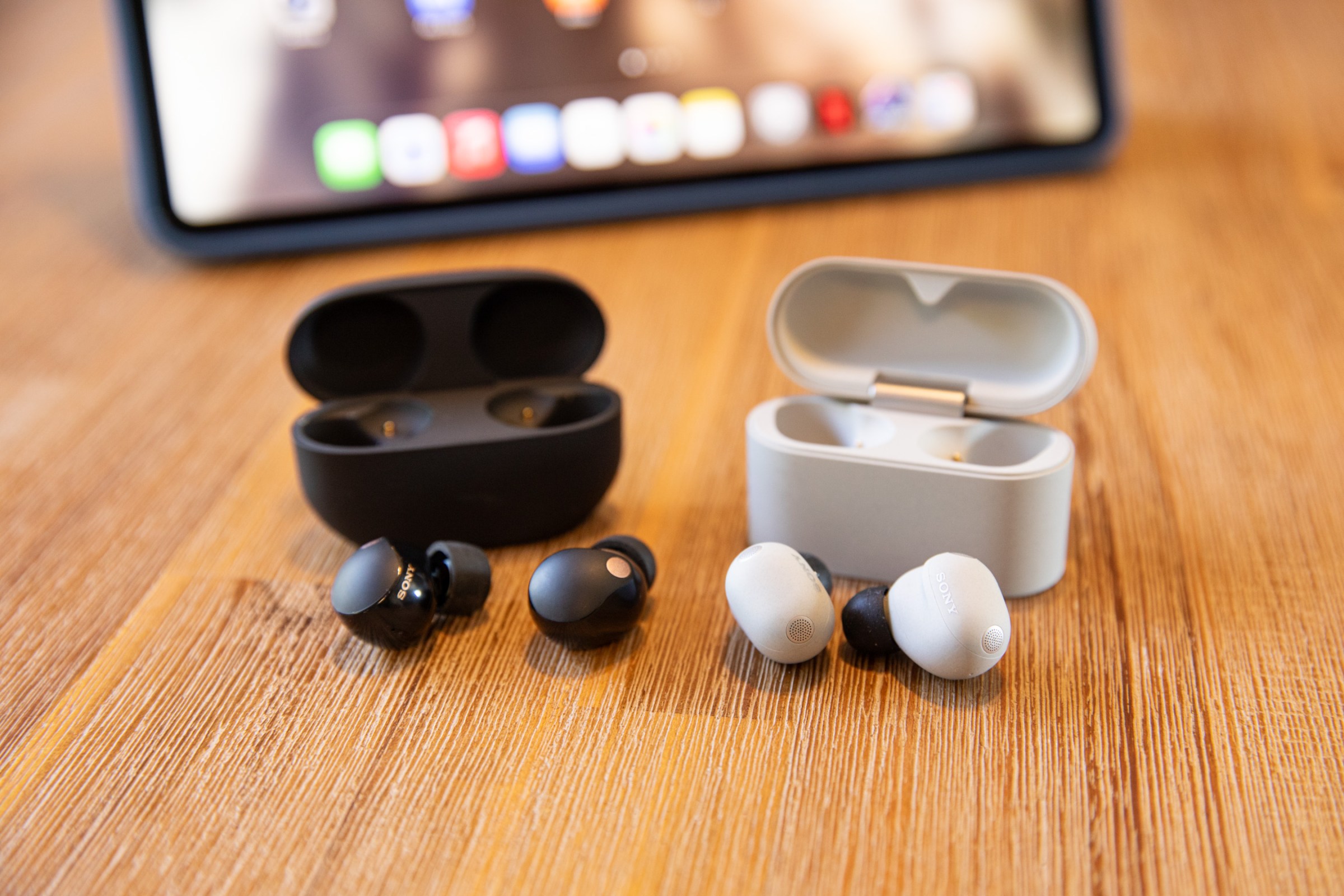 The Sony WF-1000XM5 and WF-1000XM6 earbuds and cases on a wooden table with an iPad in the background.