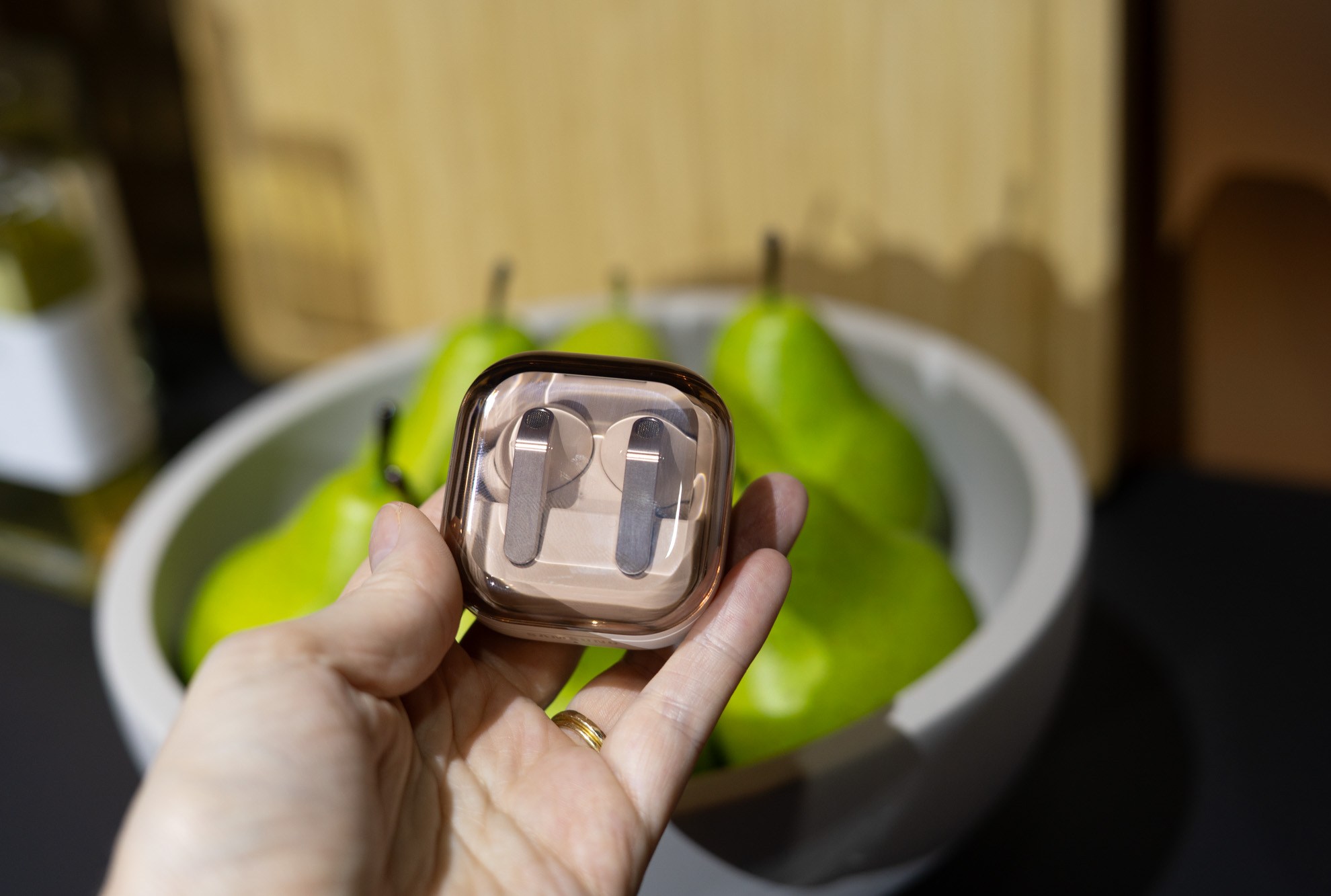A hand holding the Samsung Galaxy Buds 4 Pro in pink gold above a bowl of pears.