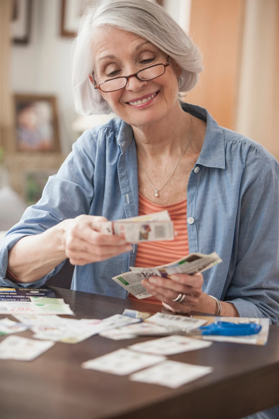 Older adult with glasses joyfully sorting coupons at a table, wearing a casual denim shirt over a striped top
