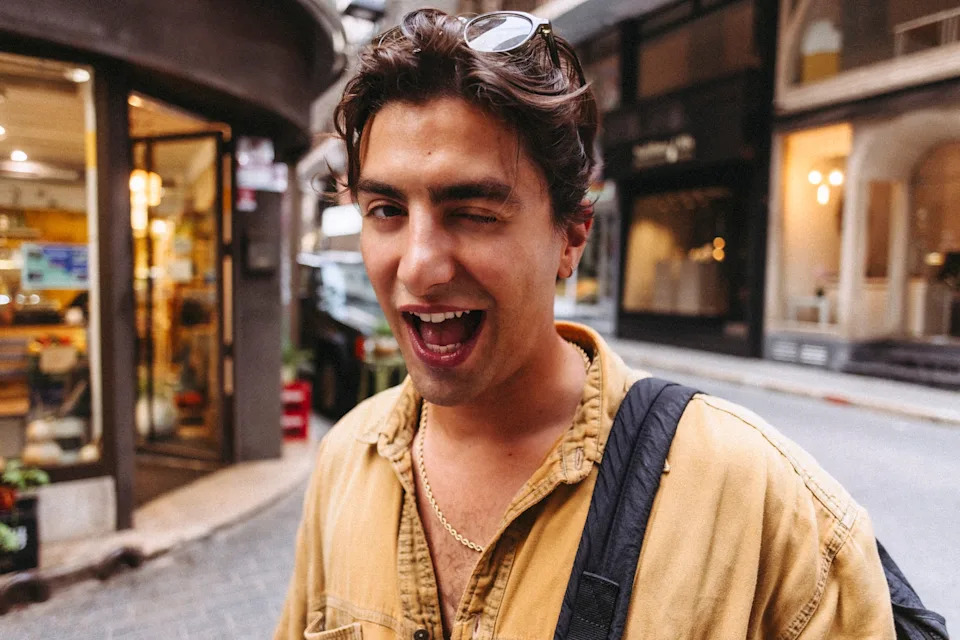 Person playfully winking at the camera on a city street, wearing casual shirt and sunglasses on head