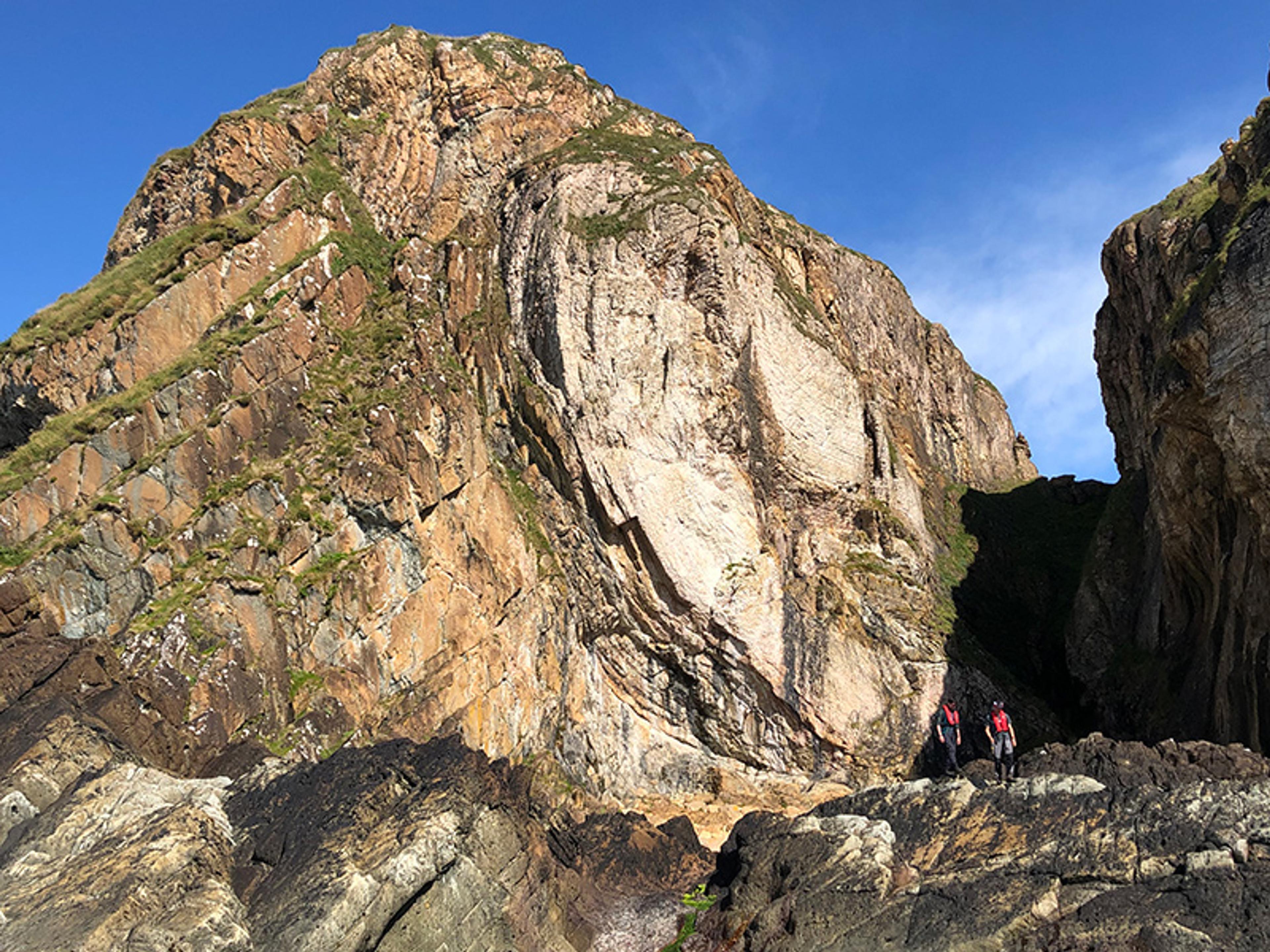 Two hikers in colourful jackets standing at the base of a large rugged rock formation under a clear blue sky.