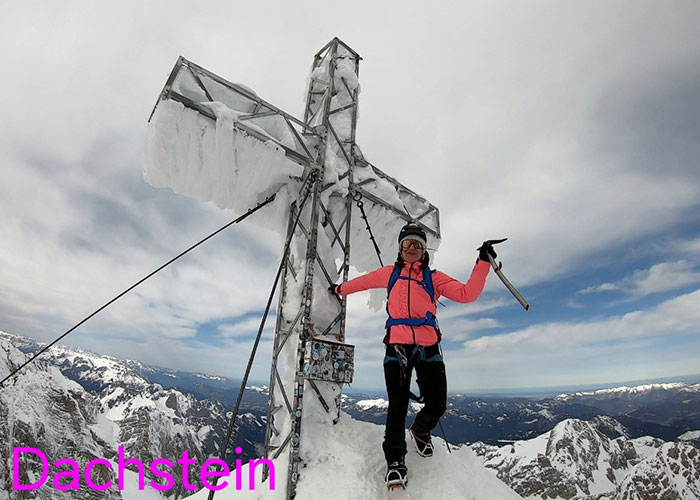 Climber standing on snowy mountain peak next to large icy cross during an alpine expedition in Dachstein.