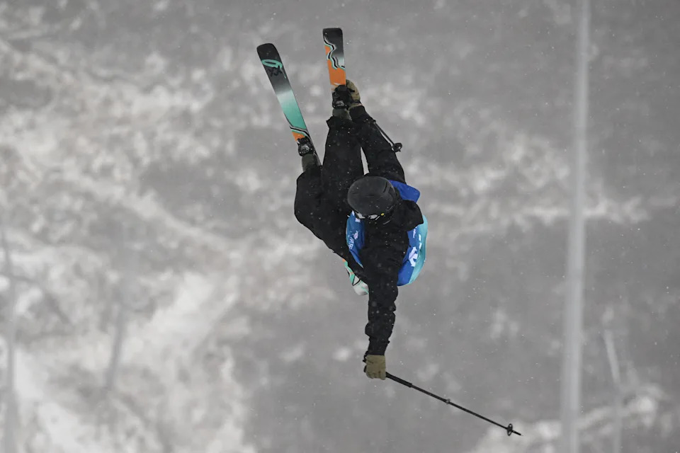 Skier performs a high-flying aerial trick with skis crossed, wearing a helmet and snow gear, against a snowy mountainous backdrop