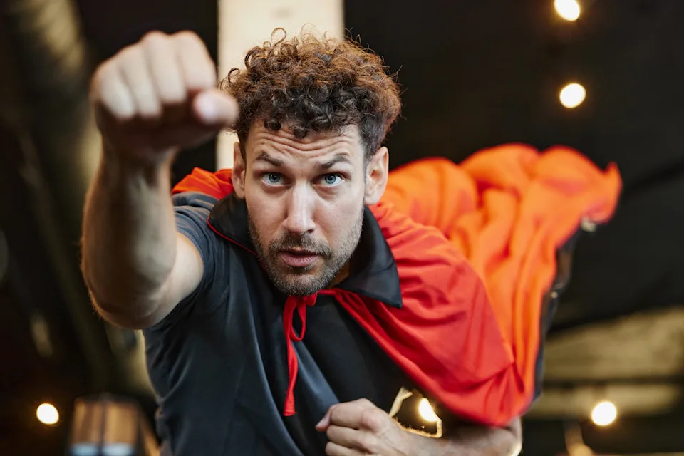 Person posing as a superhero, wearing a cape, with a focused expression, in a playful action pose indoors