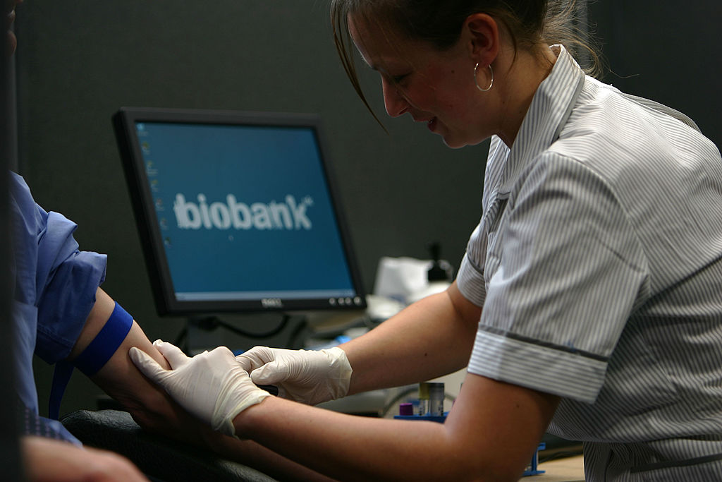 A woman with their brown hair up and wearing hoop earrings and a striped blue and white shirt uses gloved hands to stick a needle into a person's arm. The person, off camera, wears a blue shirt and a blue rubber tourniquet on their arm.