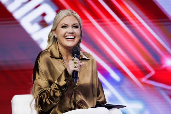 Person smiling and speaking into a microphone, wearing a satin blouse onstage with a blurred, colorful background