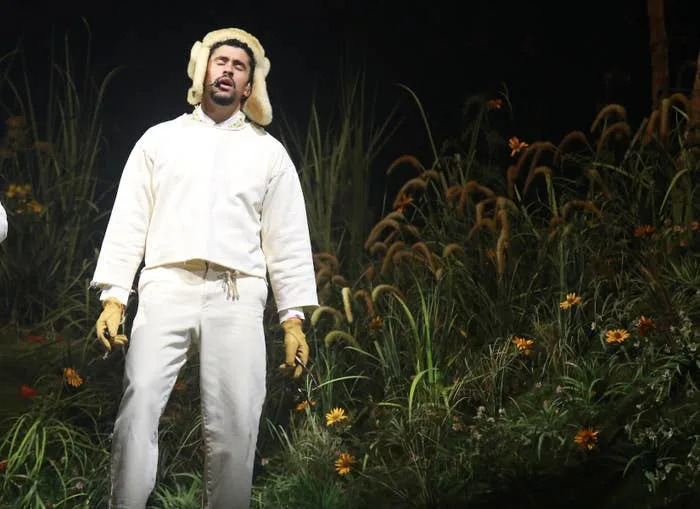 A person in a white outfit and a fluffy hat stands on stage, surrounded by tall grass and flowers, performing passionately