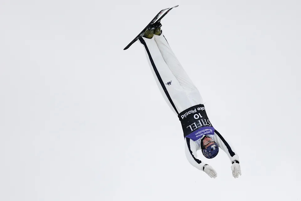 A ski jumper in mid-air, wearing a helmet and aerodynamic suit with extended arms, performs a jump against a clear sky