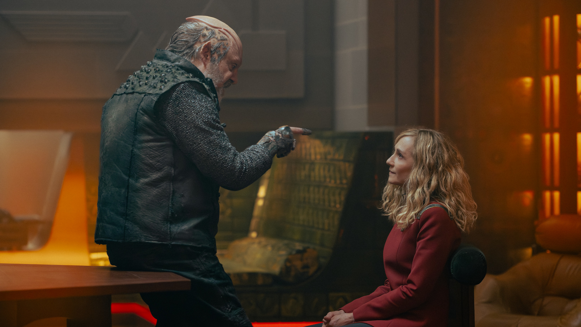 a humanoid alien with x's and o's shaved into the side of his head and a raised cranium points his finger in the face of a woman with curly blonde hair in a red officer's uniform inside a richly appointed office
