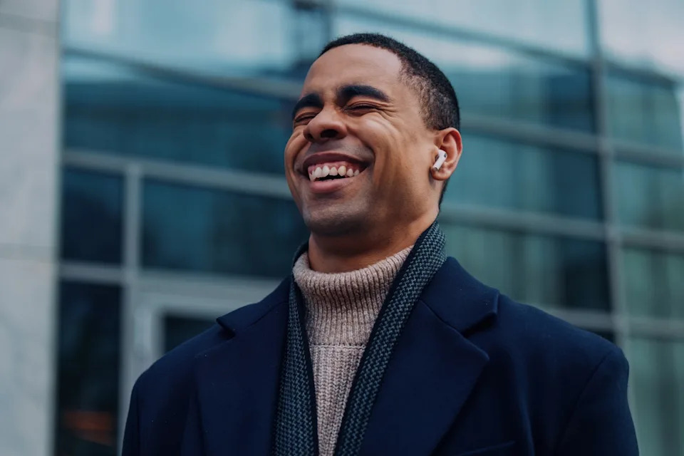 Man outdoors, smiling with eyes closed, wearing earbuds, turtleneck sweater, scarf, and coat. Standing in front of glass building