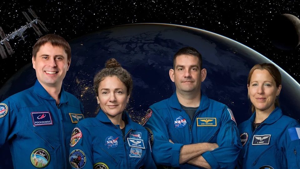 four humans stand in blue jumpsuits in front of a background of a dark earth and shadowed moon, with a small ISS in the top left. 